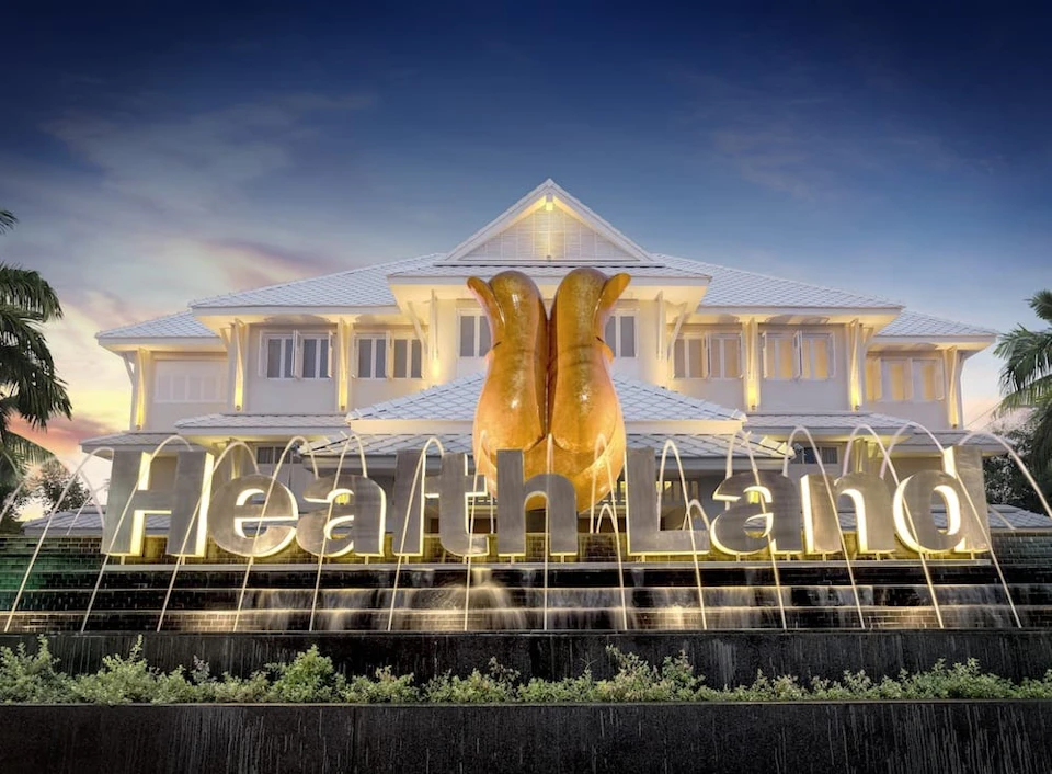 A large building with lit signage reading 'Health Land' in front of a sculpture, set against a twilight sky.