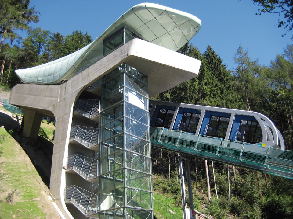 A modern funicular railway with glass-walled elevator and staircase structure, surrounded by trees and a clear blue sky.