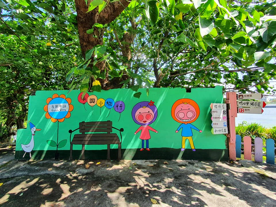 A colorful painted wall under a tree, featuring cartoon characters, a bench, flowers, and balloons with Chinese text.