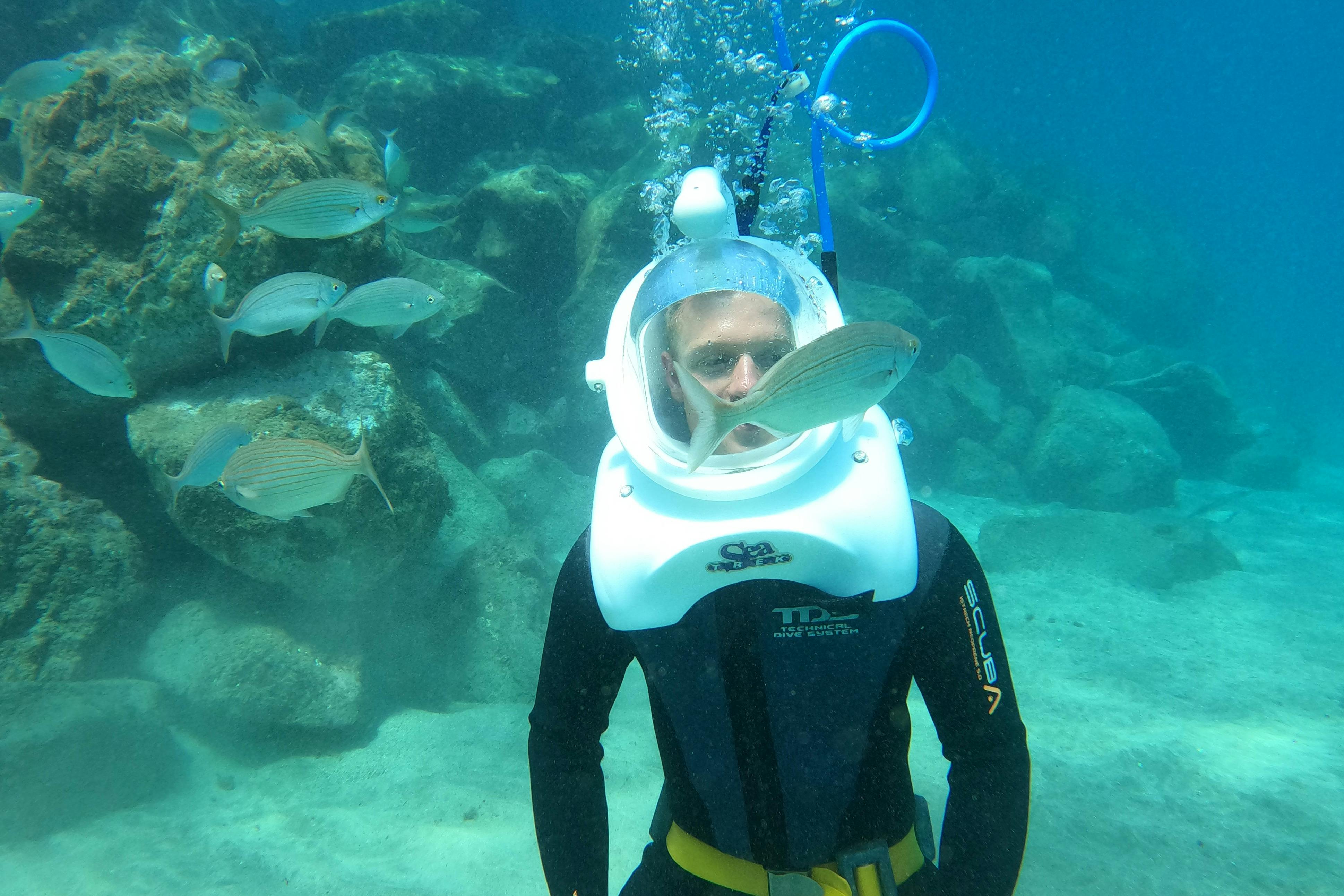 Person underwater wearing a helmet dive suit, surrounded by fish, with bubbles rising from the helmet. Rocky background visible.