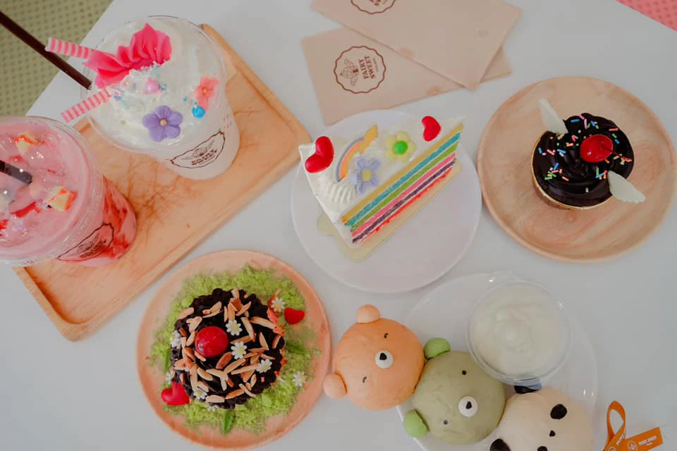 A selection of colorful desserts on plates, including a rainbow cake, cupcakes, and decorated pastries on a table.