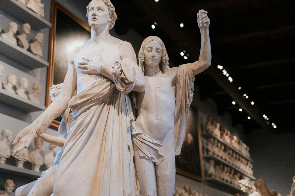Two marble statues displayed in an art gallery with numerous other sculptures visible in the background.