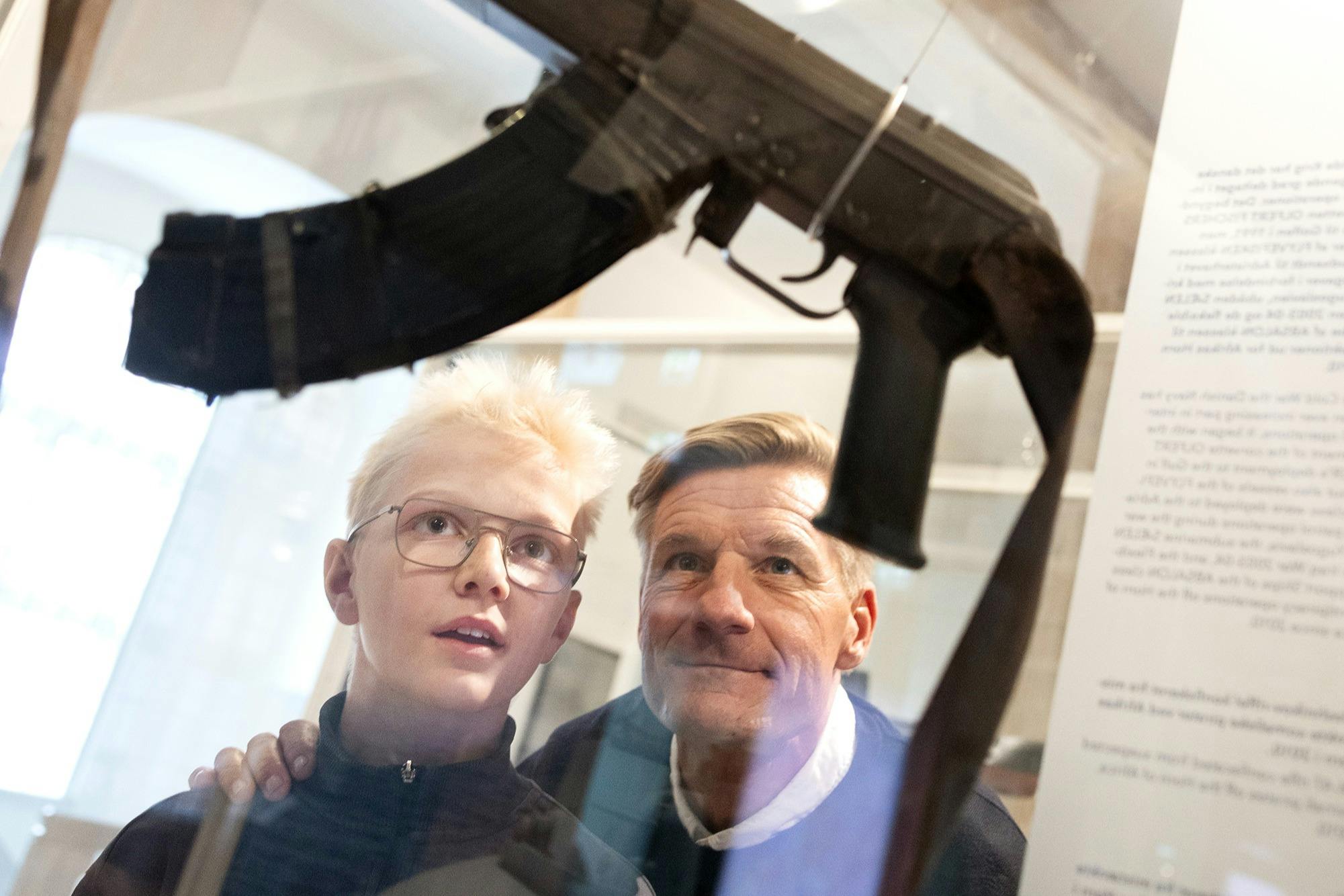 Et barn og en voksen ser på et skydevåben, der vises på et museum, med fokus på deres udtryk og udstillingen.