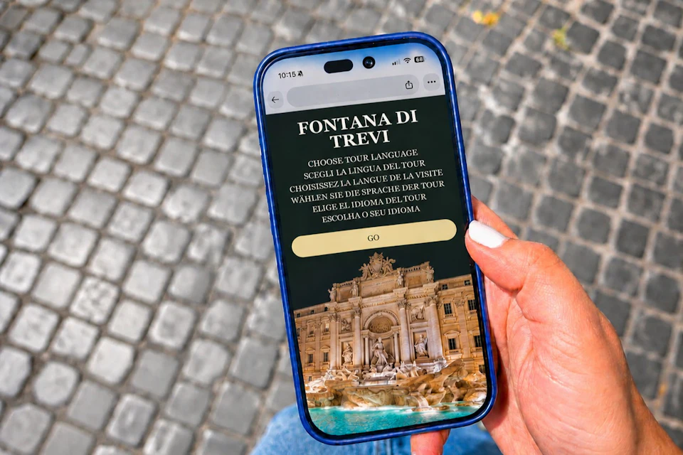 A hand holding a smartphone displaying a tour selection screen for the Trevi Fountain, with a cobblestone ground in the background.