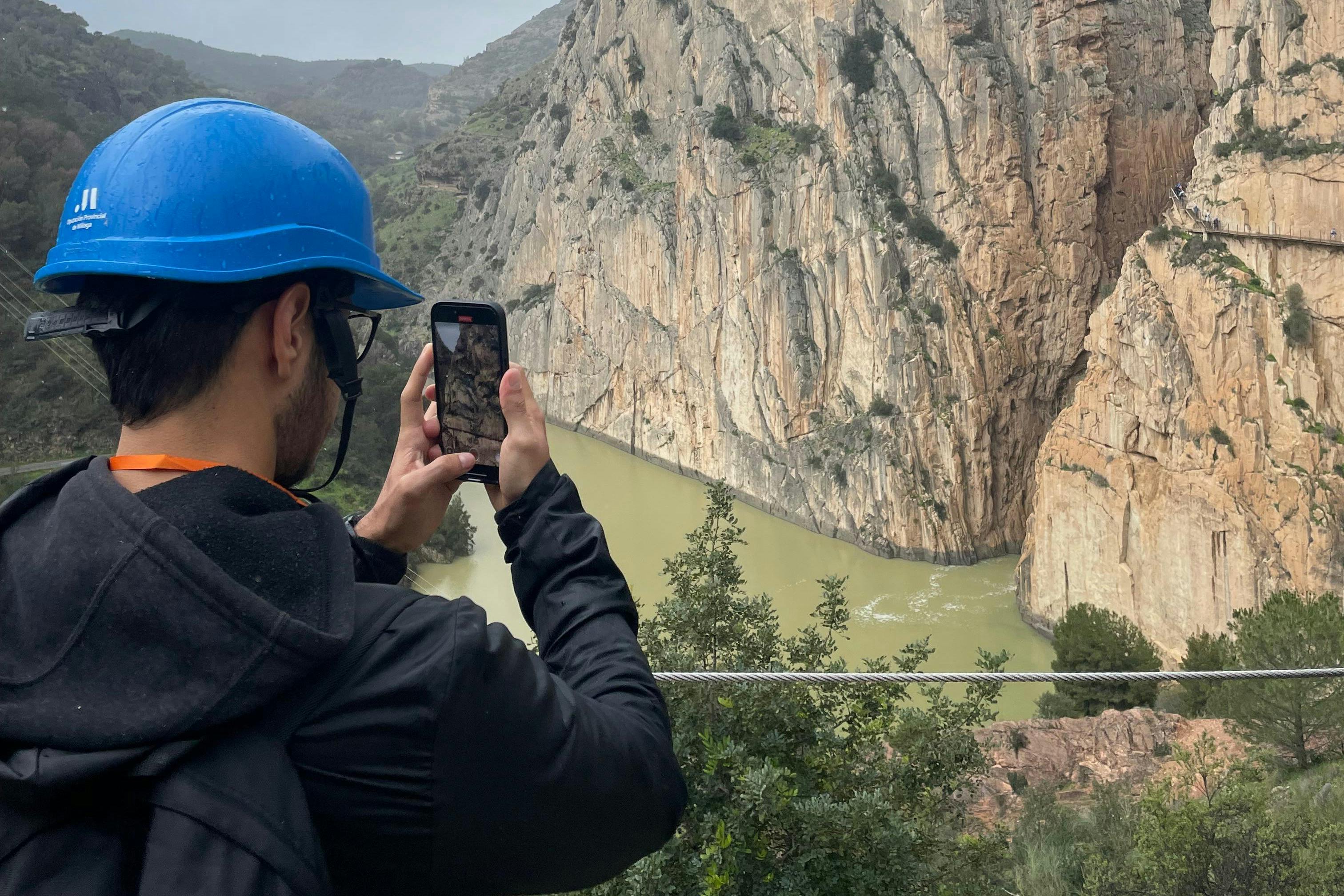 A person wearing a blue helmet photographs a steep canyon with a river flowing through it using a smartphone.