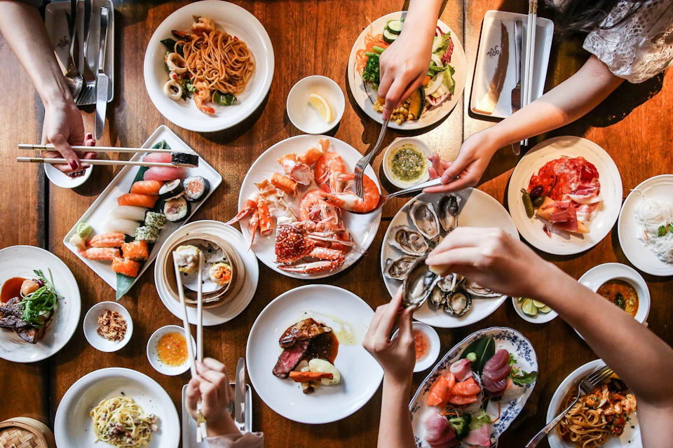 A table with various seafood and sushi dishes, surrounded by hands reaching for food with utensils and chopsticks.
