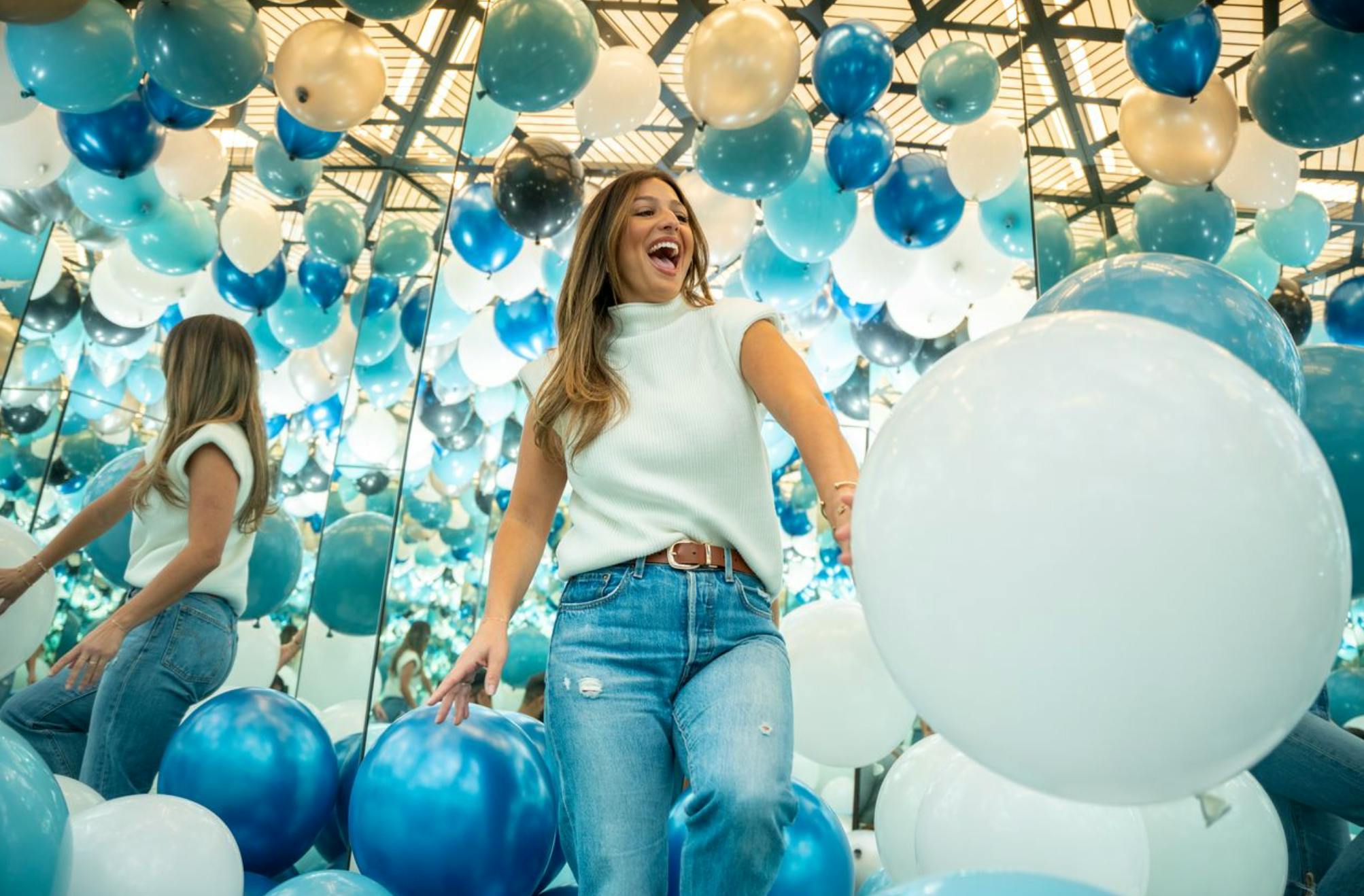 En kvinna i avslappnad klädsel ler glatt i ett rum fyllt av blå och vita ballonger och spegelreflektioner.