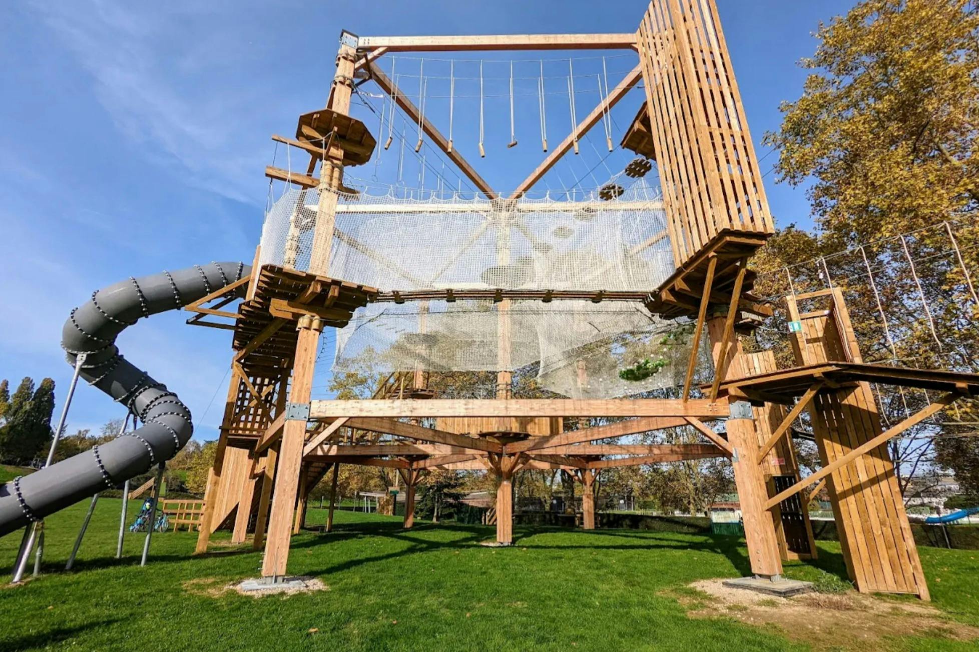 En utomhus äventyrsbana med träkonstruktioner, nät, en spiralrutschbana och grönska under en klarblå himmel.