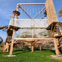 En utomhus äventyrsbana med träkonstruktioner, nät, en spiralrutschbana och grönska under en klarblå himmel.