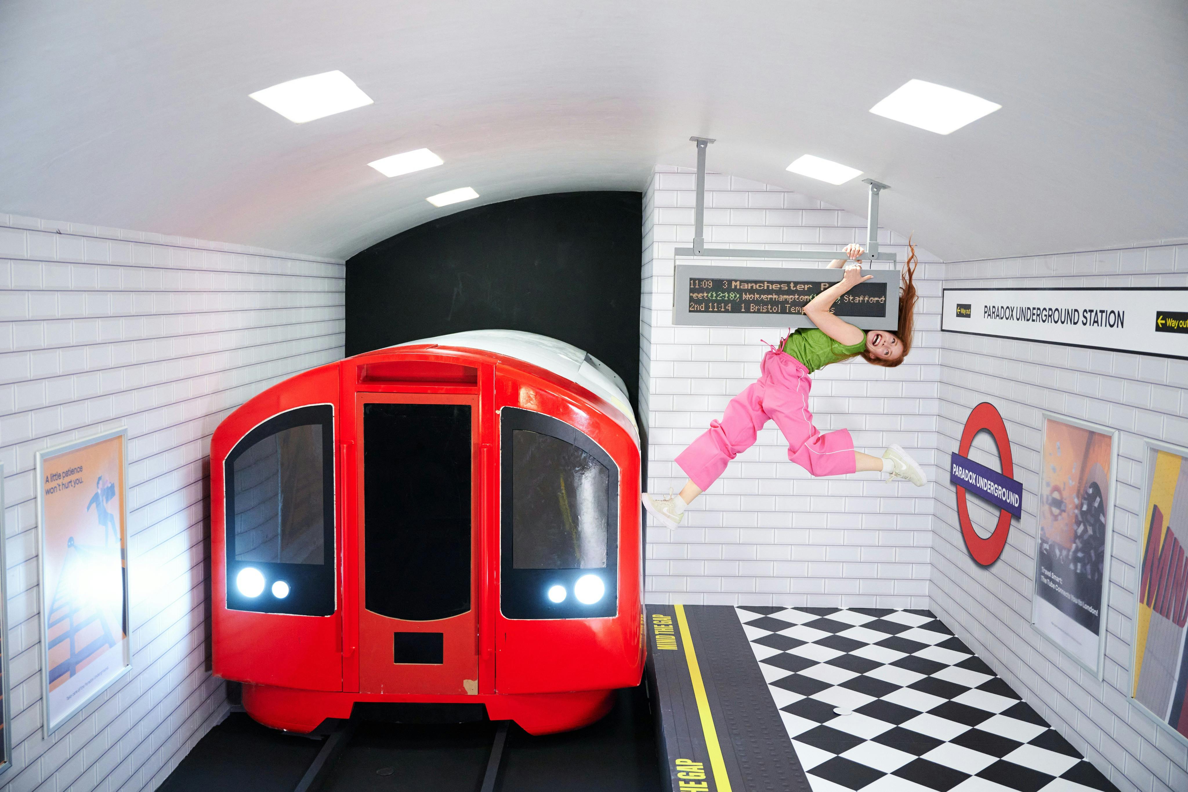 Člověk v růžových kalhotách a zelené košili visí vodorovně z cedule uvnitř makety stanice metra s červeným vlakem.