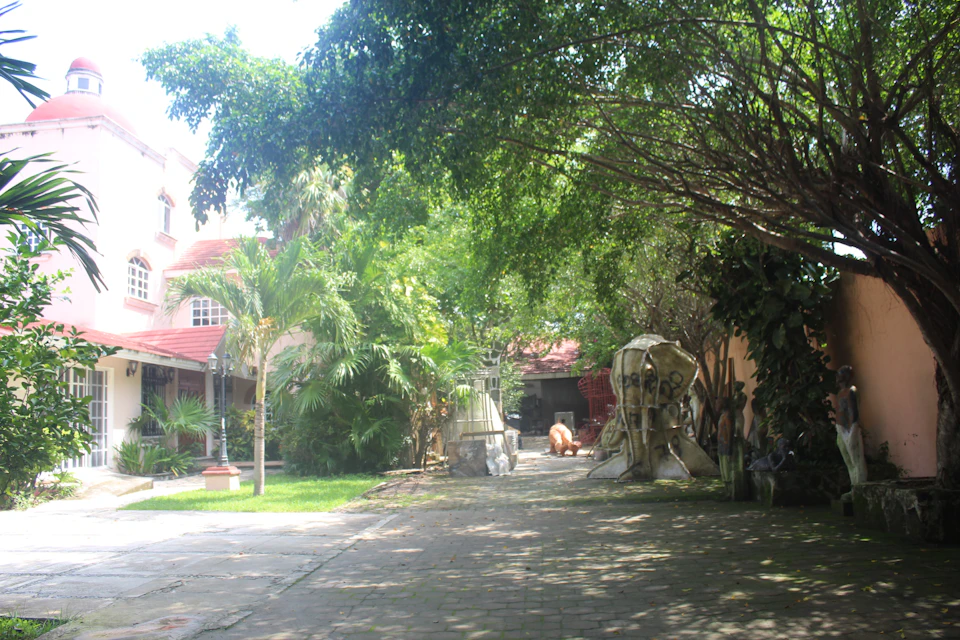 A sunlit, tree-lined courtyard with sculptures, a red-roofed building, and lush greenery.