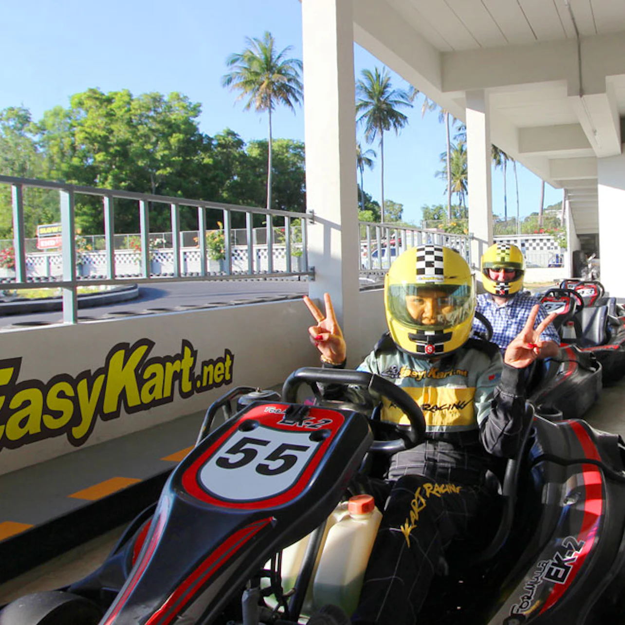 Two people in go-karts, wearing helmets, preparing to race at an outdoor karting track with palm trees in the background.