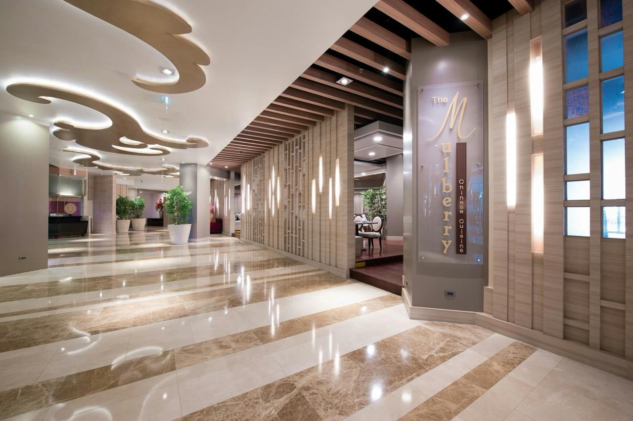 A stylish hotel lobby with marble floors, modern decor, potted plants, and a sign reading "The Mulberry" on the wall.