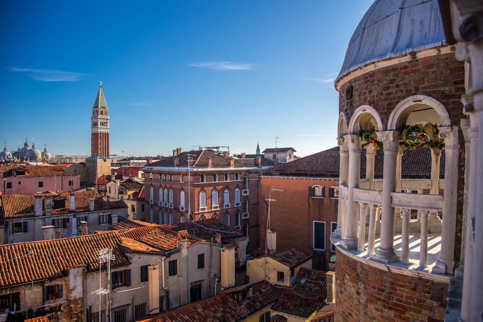 Rooftop view of Venetian buildings with red tiles, the bell tower of St. Mark's Basilica, and a clear blue sky.