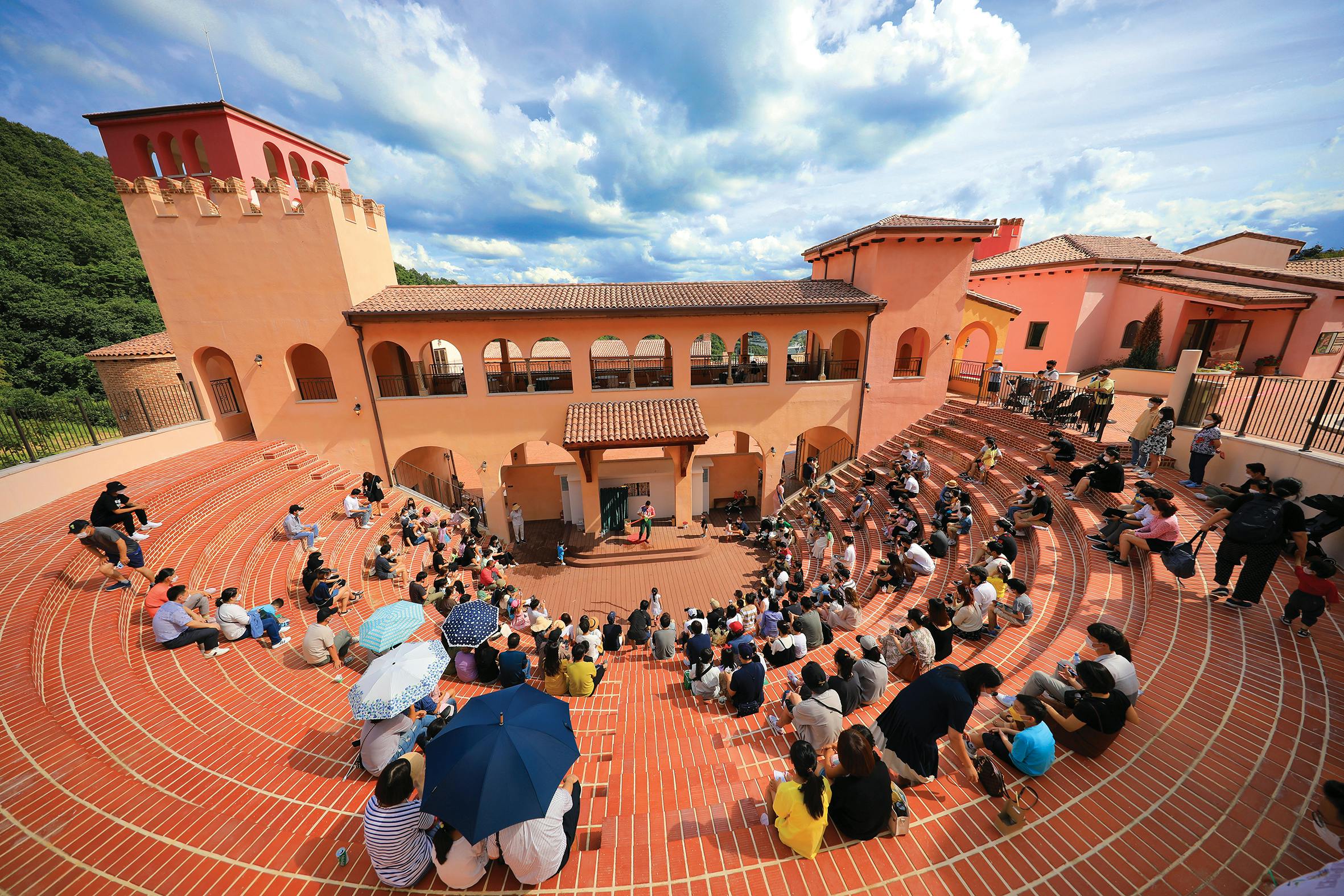 An outdoor amphitheater with terracotta steps, a central stage, and an audience gathered under a partly cloudy sky.