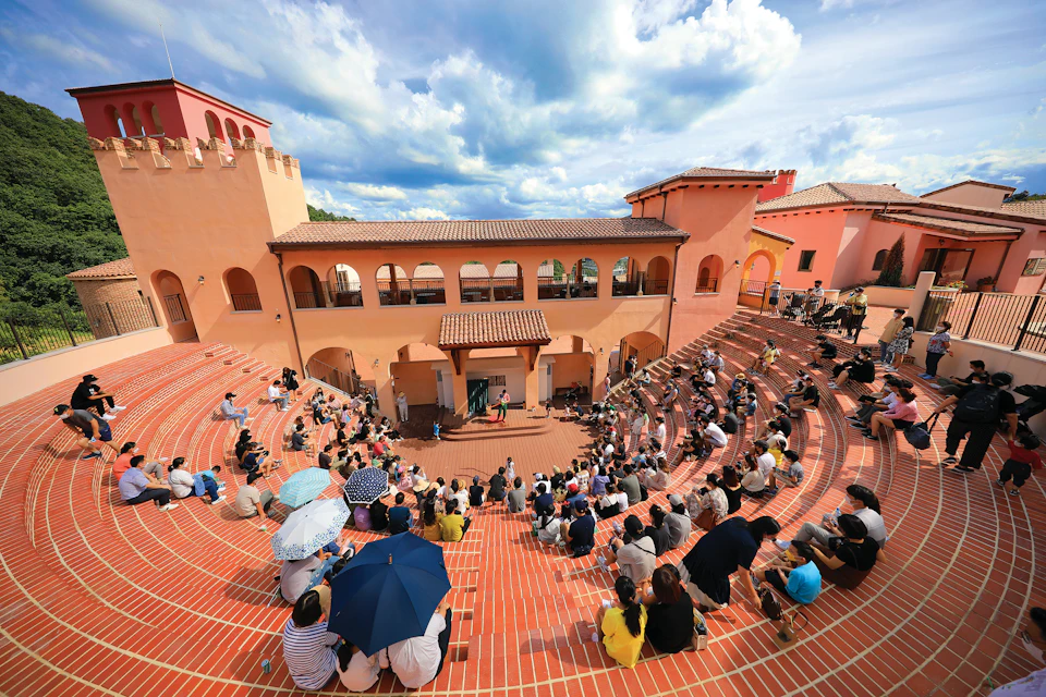 An outdoor amphitheater with terracotta steps, a central stage, and an audience gathered under a partly cloudy sky.