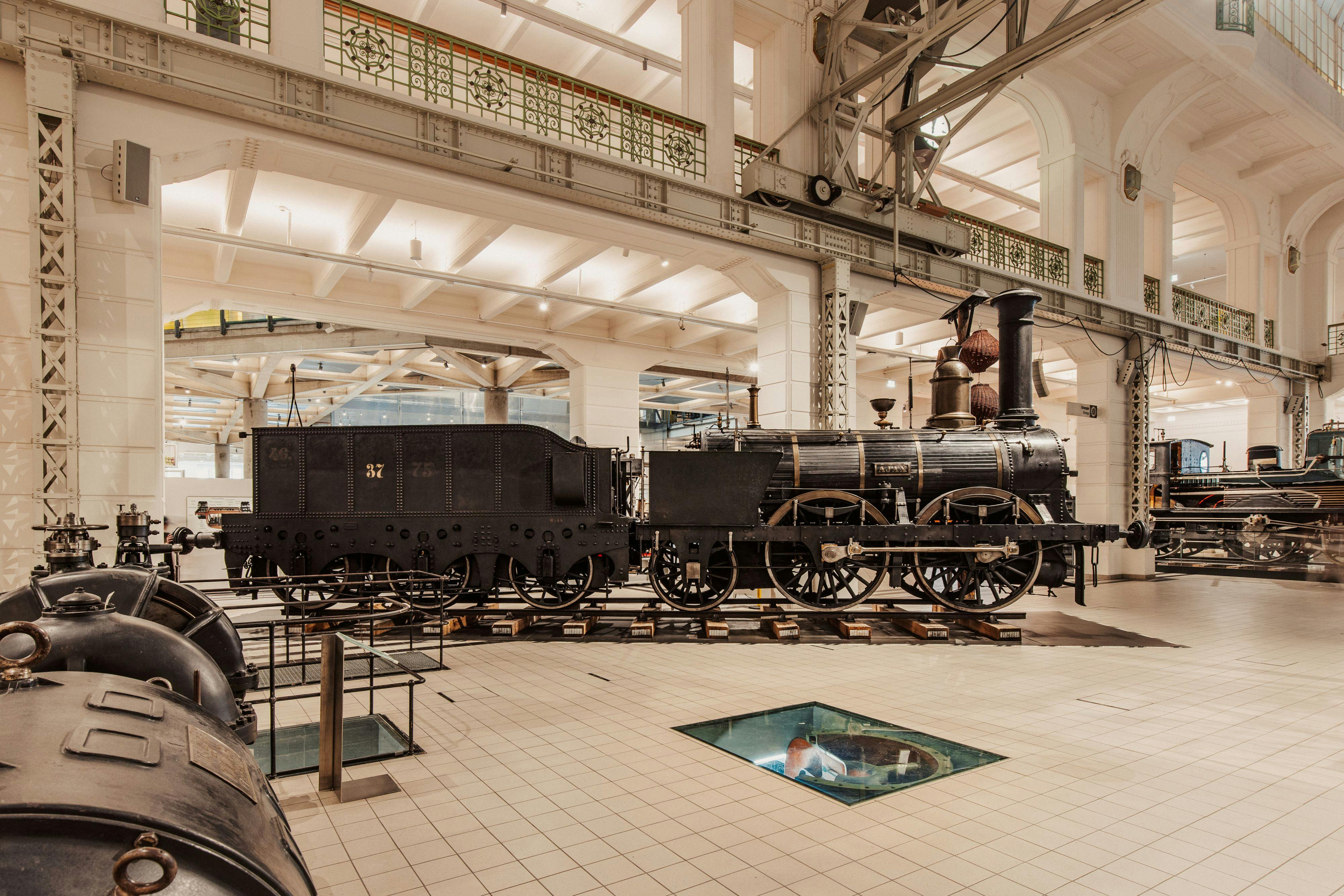 Historická parní lokomotiva v dobře osvětleném prostorném muzeu s architektonickými detaily a skleněnou podlahovou deskou.