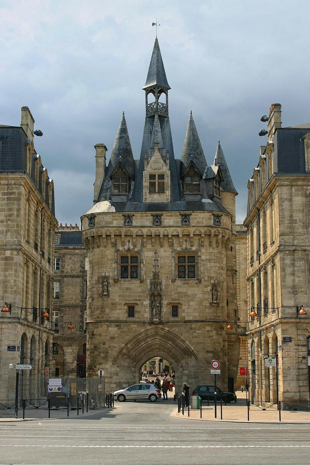 Een historisch stenen poortgebouw met puntige spitsen, geflankeerd door twee gebouwen, en een paar mensen en auto's die door de poort rijden.