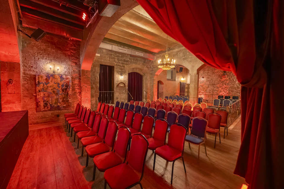 A dimly lit theater with red and blue chairs arranged in rows, stone walls, chandeliers, and decorative curtains.