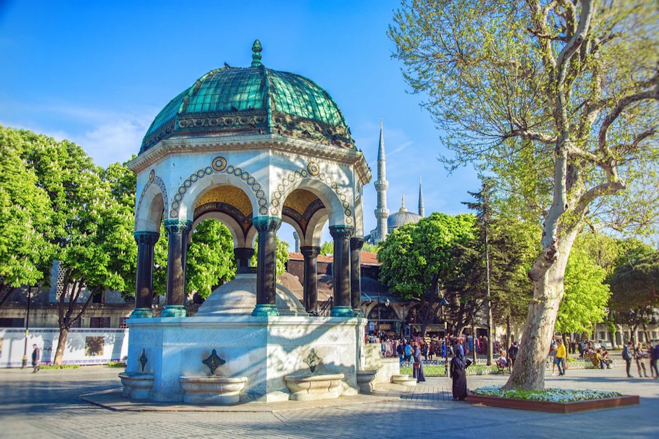 A decorative fountain with a green dome in a park setting, with trees, people, and a mosque in the background.