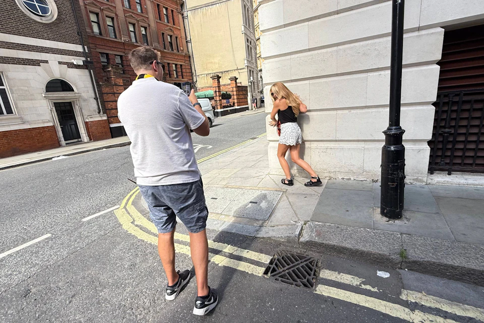 A man photographing a woman posing against the corner of a building on a city street during daylight.