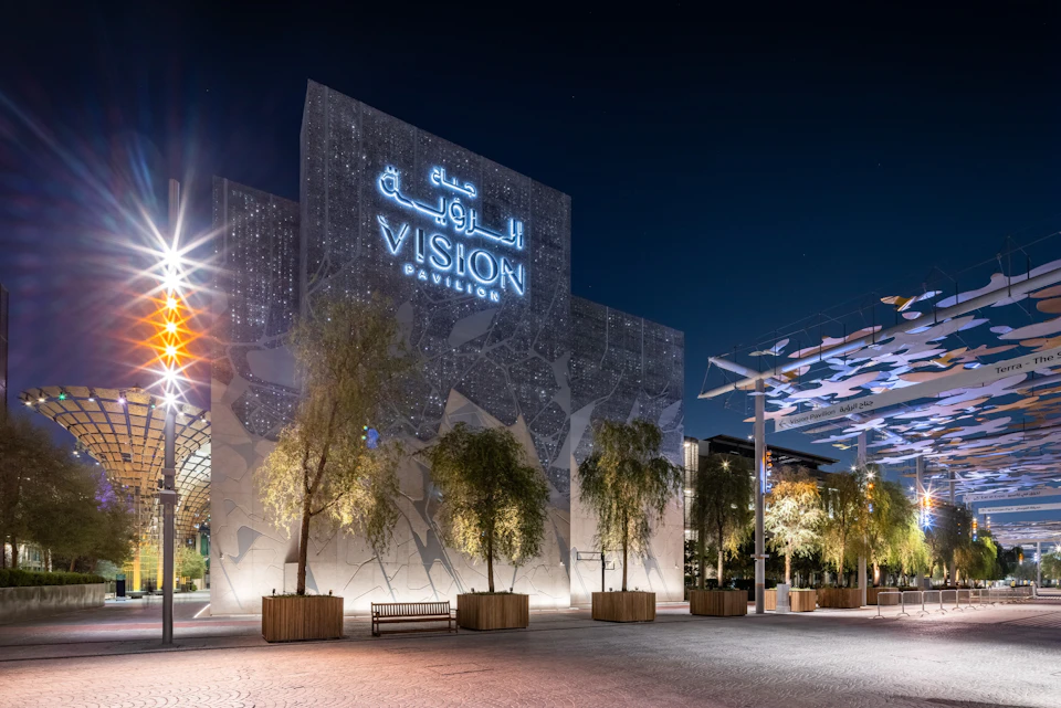 A modern, illuminated building at night labeled 'Vision Pavilion,' surrounded by trees and benches in a plaza.