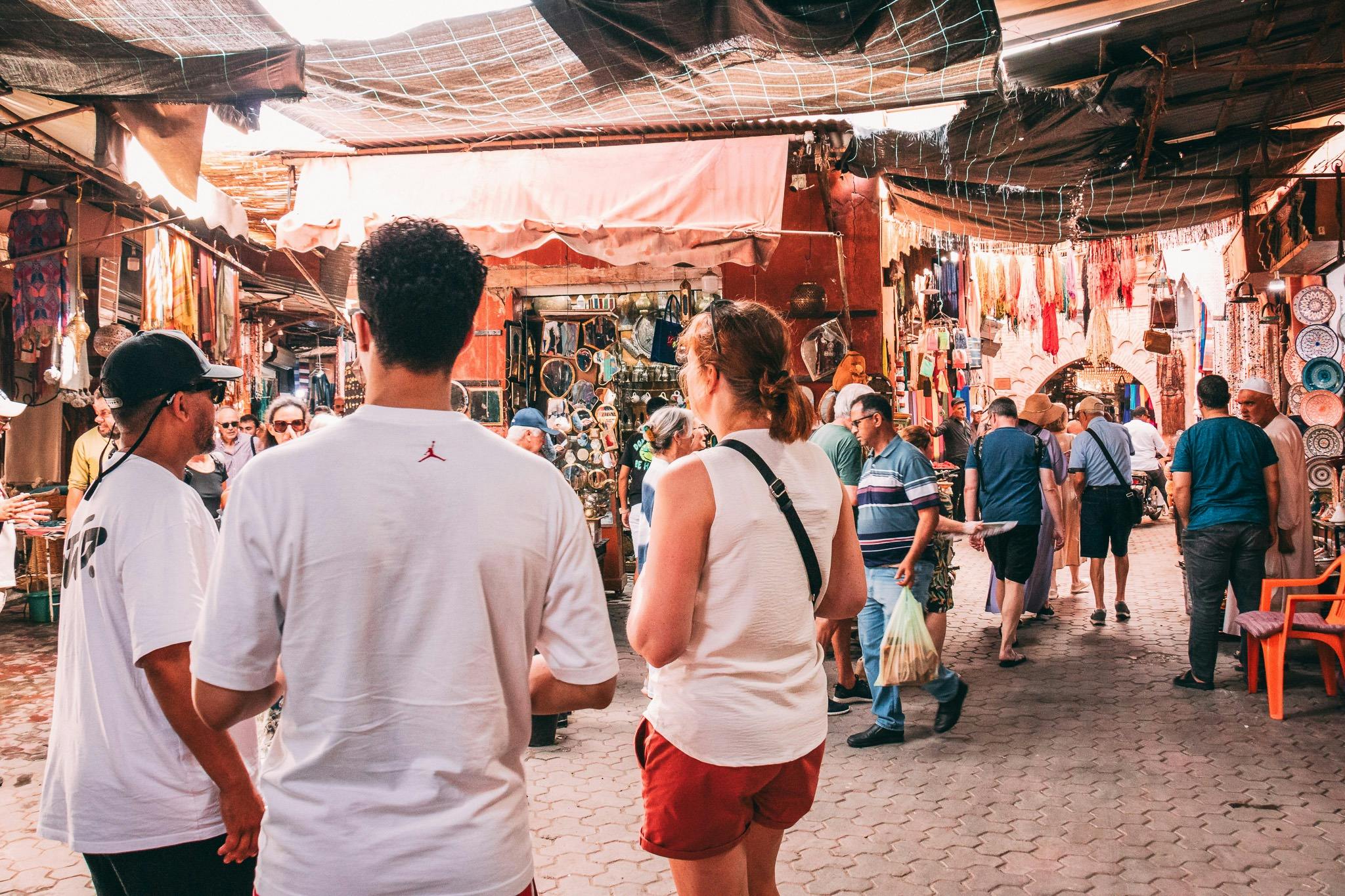 Trois personnes traversent un marché animé, avec des étals de marchandises et de décorations colorées, sous un auvent de bâches.