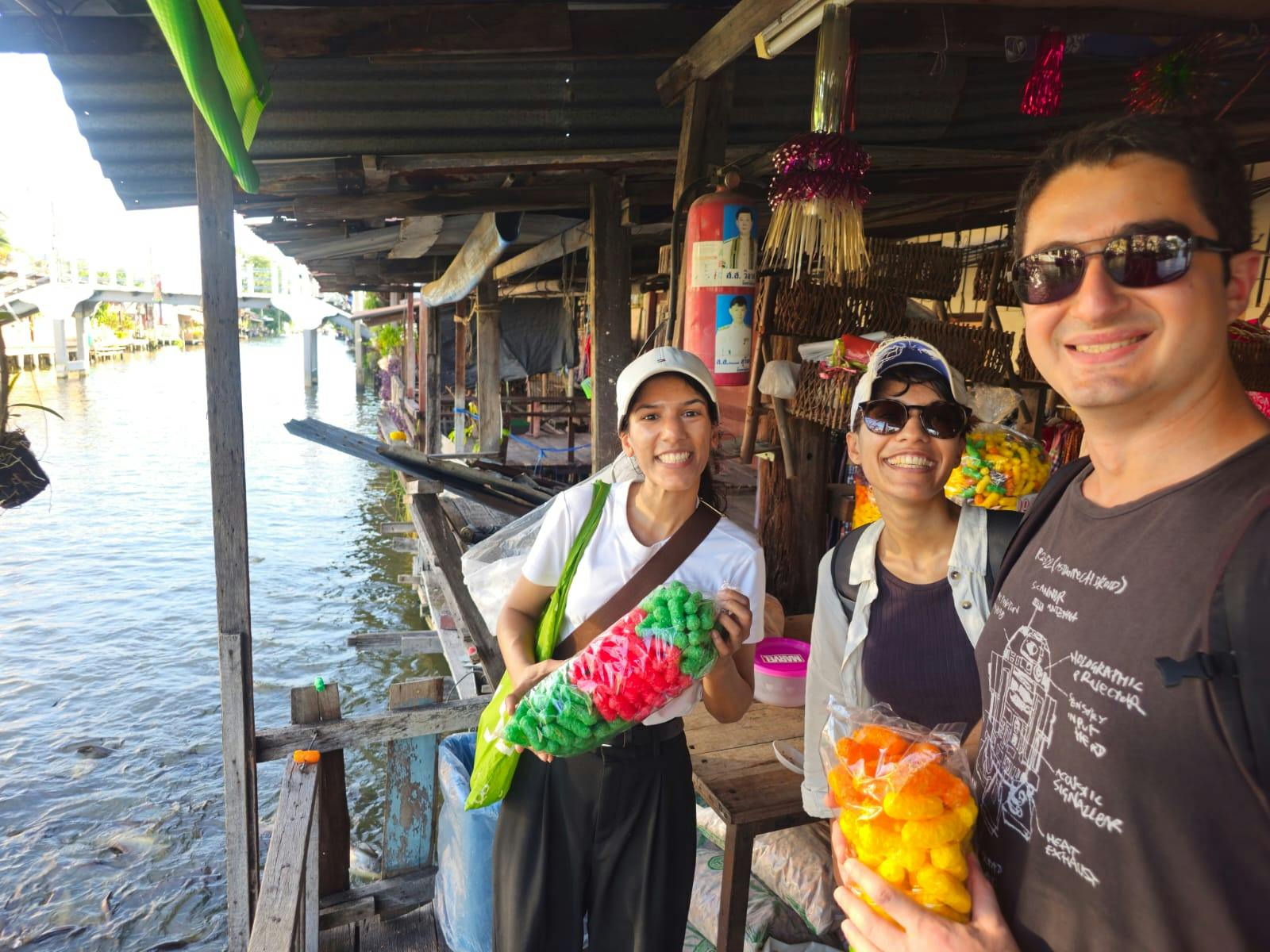 Tres persones somrient i amb bosses d'aperitius de colors en un mercat a l'aire lliure al costat del mar. Les parades protegides són visibles darrere d'ells.