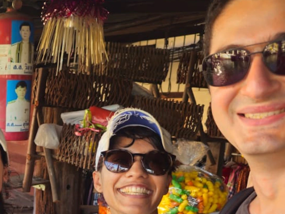 Three people smiling and holding bags of colorful snacks at an outdoor market by a waterfront. Sheltered stalls are visible behind them.