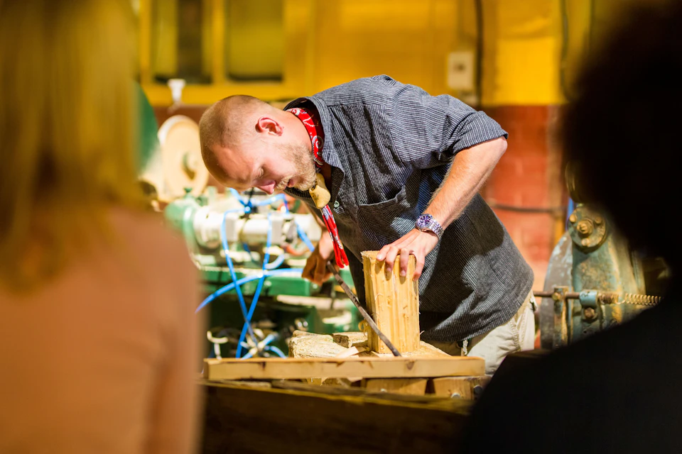 A man in a workshop using a hand plane on a piece of wood, surrounded by various tools and machinery.
