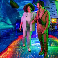 Un uomo e una donna che camminano su un pavimento illuminato e colorato, sorridendosi l'un l'altro, in un ambiente interno vivace.