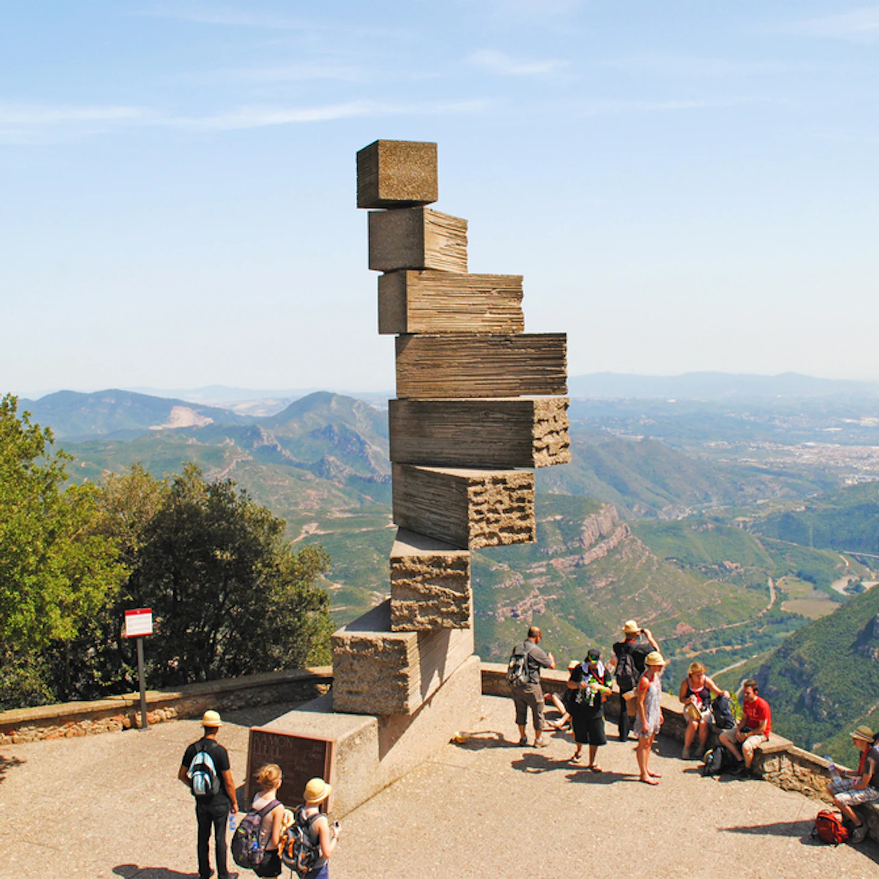 Tourists gather around a large, abstract stone sculpture on a viewing platform overlooking a mountainous landscape.