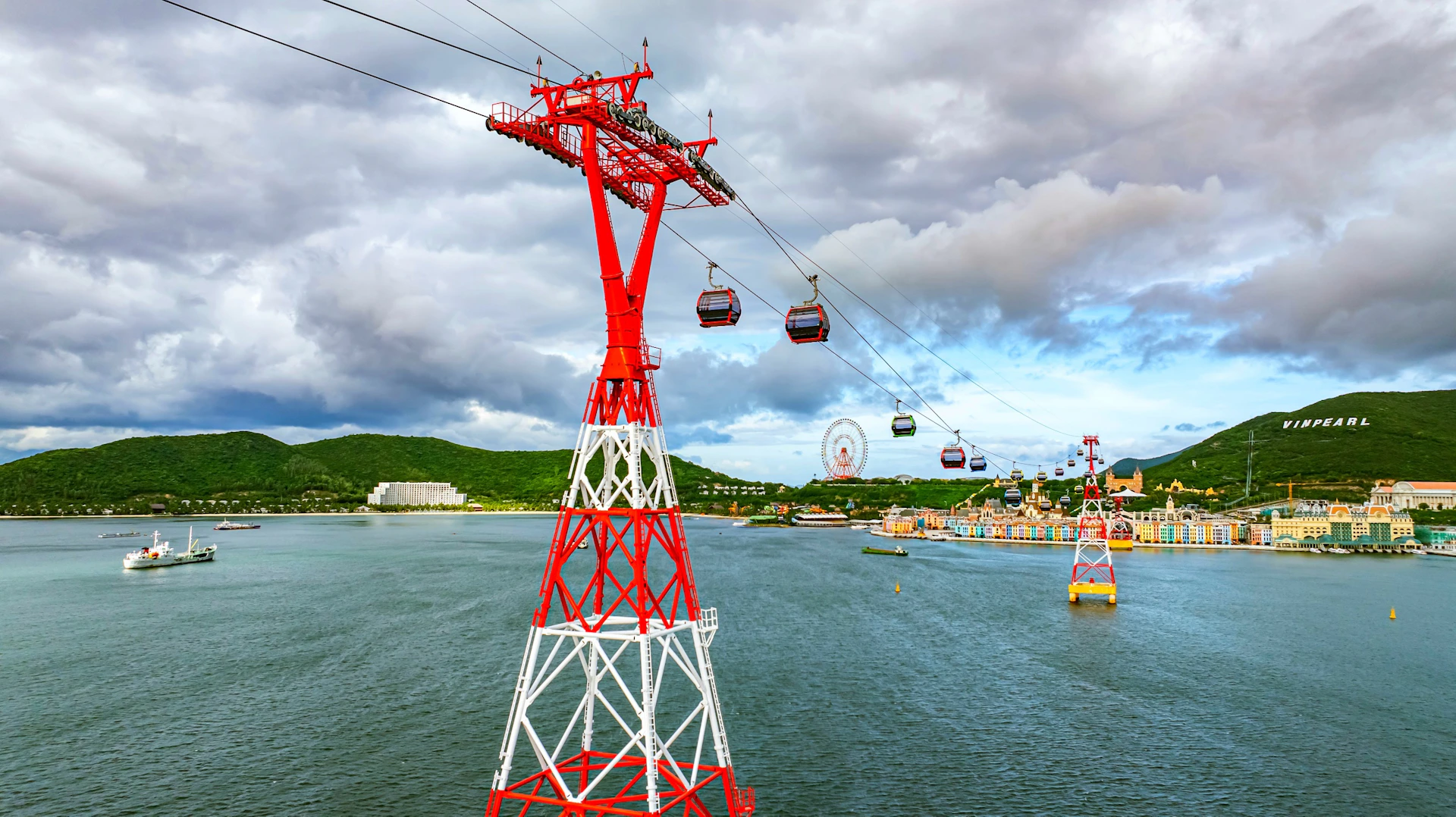 A red and white cable car tower over water with gondolas, a distant ferris wheel, and a colorful shoreline under cloudy skies.
