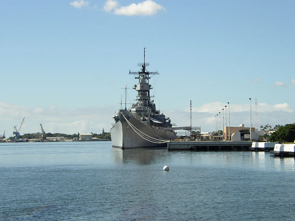 Battleship docked at a harbor with a clear blue sky, calm water, and visible buildings in the background.