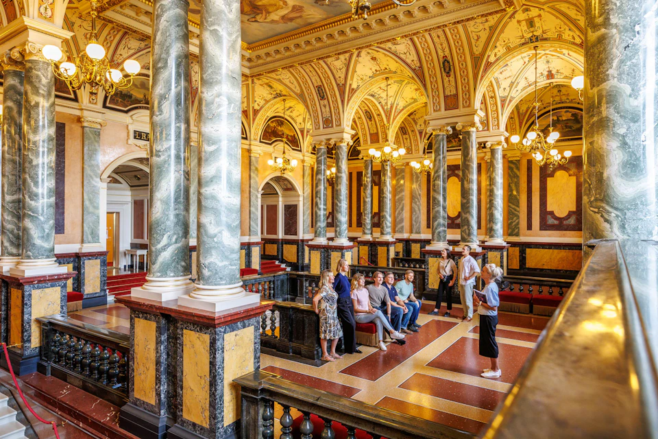 A group of people in a grand interior with ornate columns, arched ceilings, and decorative lighting.