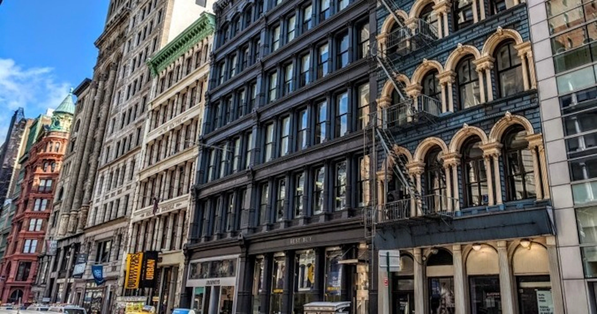 A street view showing historic multi-story buildings with dark facades and ornate fire escapes in an urban setting.