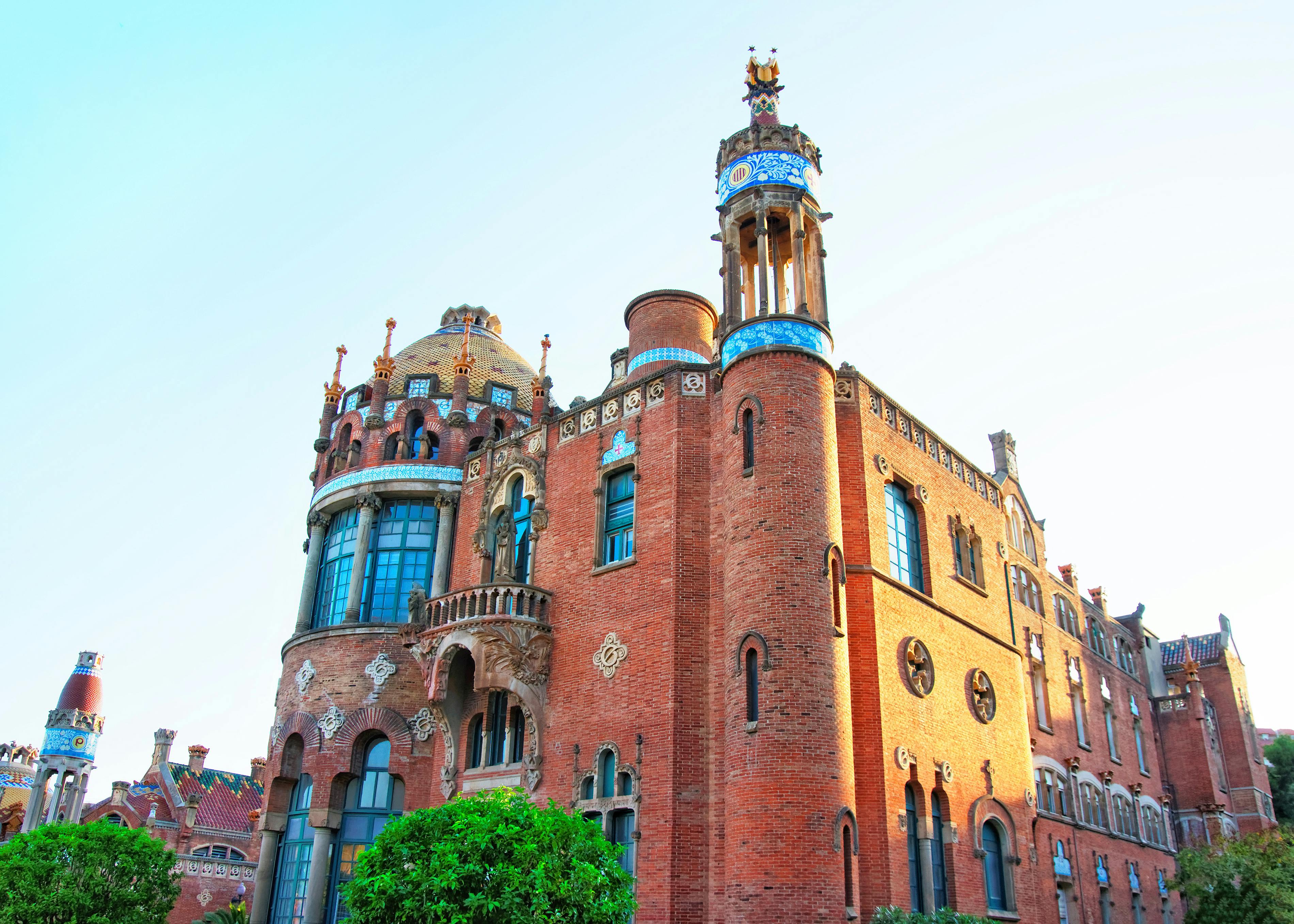 Red brick building with rounded towers, decorative elements, and arched windows, set against a clear sky. Green bushes in front.