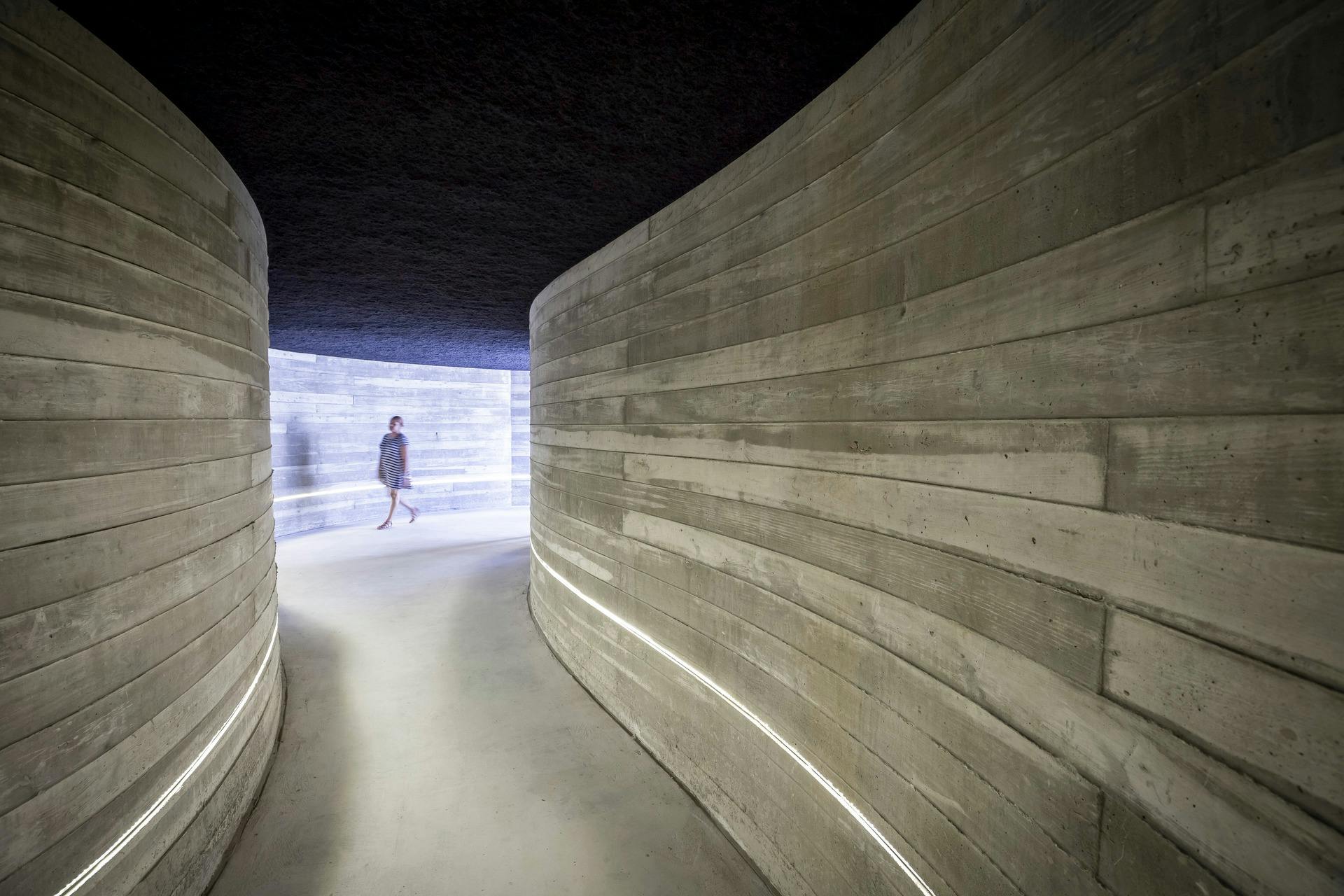 한 사람이 하단 가장자리를 따라 밝은 선이 있는 어두운 조명의 곡선형 콘크리트 복도를 걷고 있습니다.