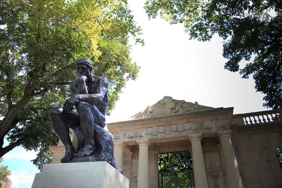 Bronze statue of a man deep in thought in front of a neoclassical building with columns and a pediment, surrounded by trees.
