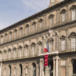 En historisk byggnad med välvda fönster och pelare, prydd med italienska och EU-flaggor, och statyer vid ingången.