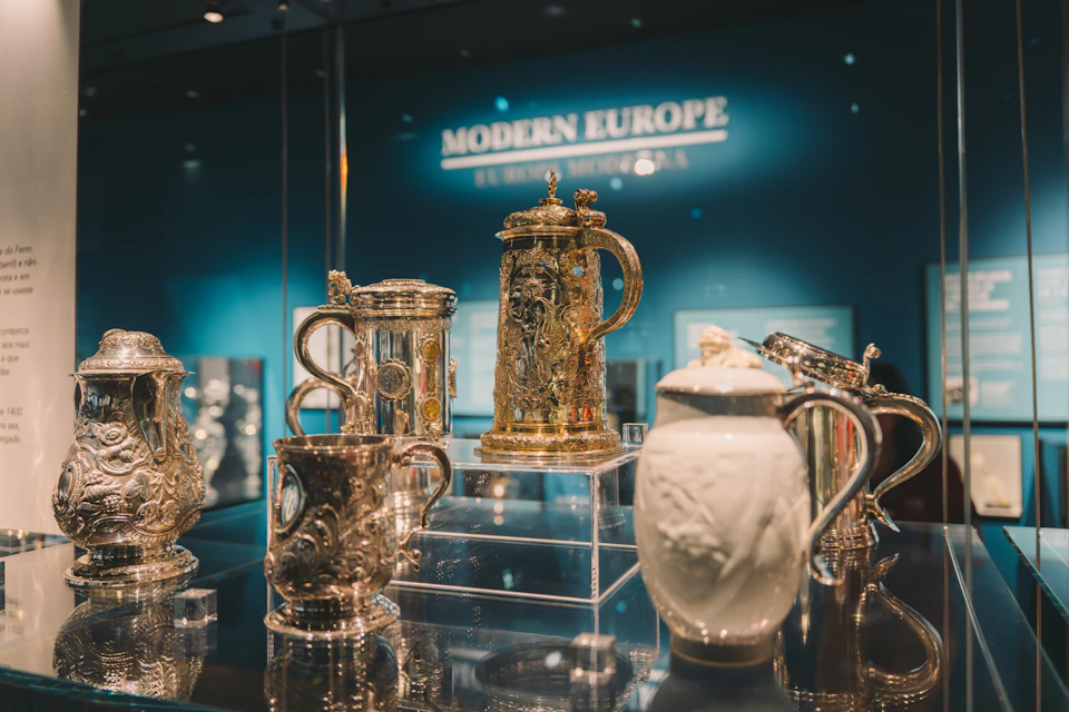 A museum display of ornate silver and ceramic vessels under the label 'Modern Europe' in a well-lit exhibition.