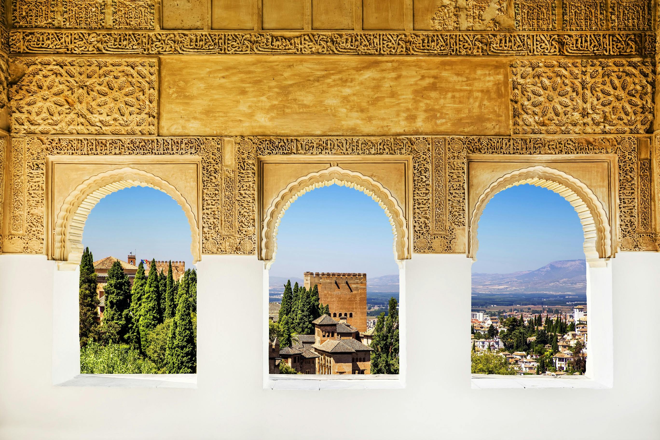 Three intricately designed arches frame a view of trees, historical buildings, and distant mountains under a clear blue sky.