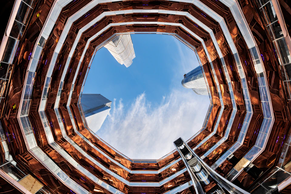 Upward view through an octagonal structure with copper-colored lining, revealing skyscrapers and blue sky with scattered clouds.
