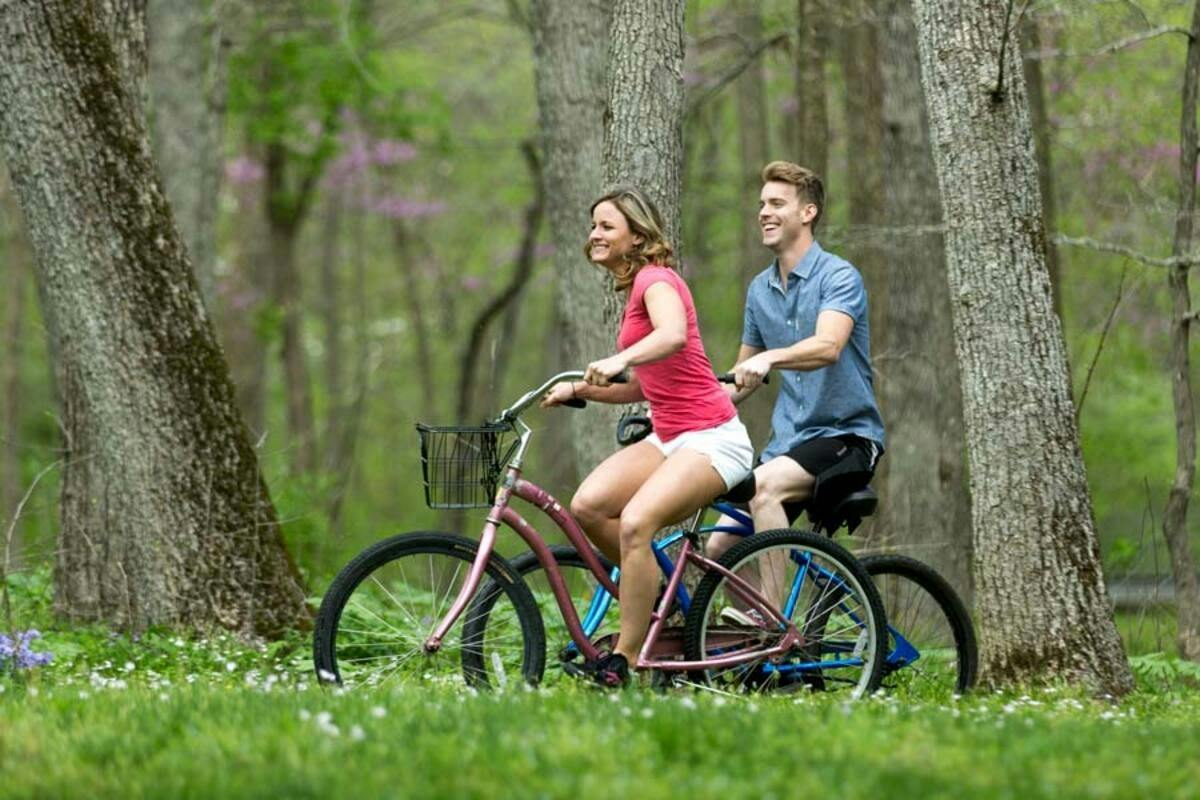 A man and a woman riding bicycles on a grassy path in a wooded area, smiling and surrounded by trees.