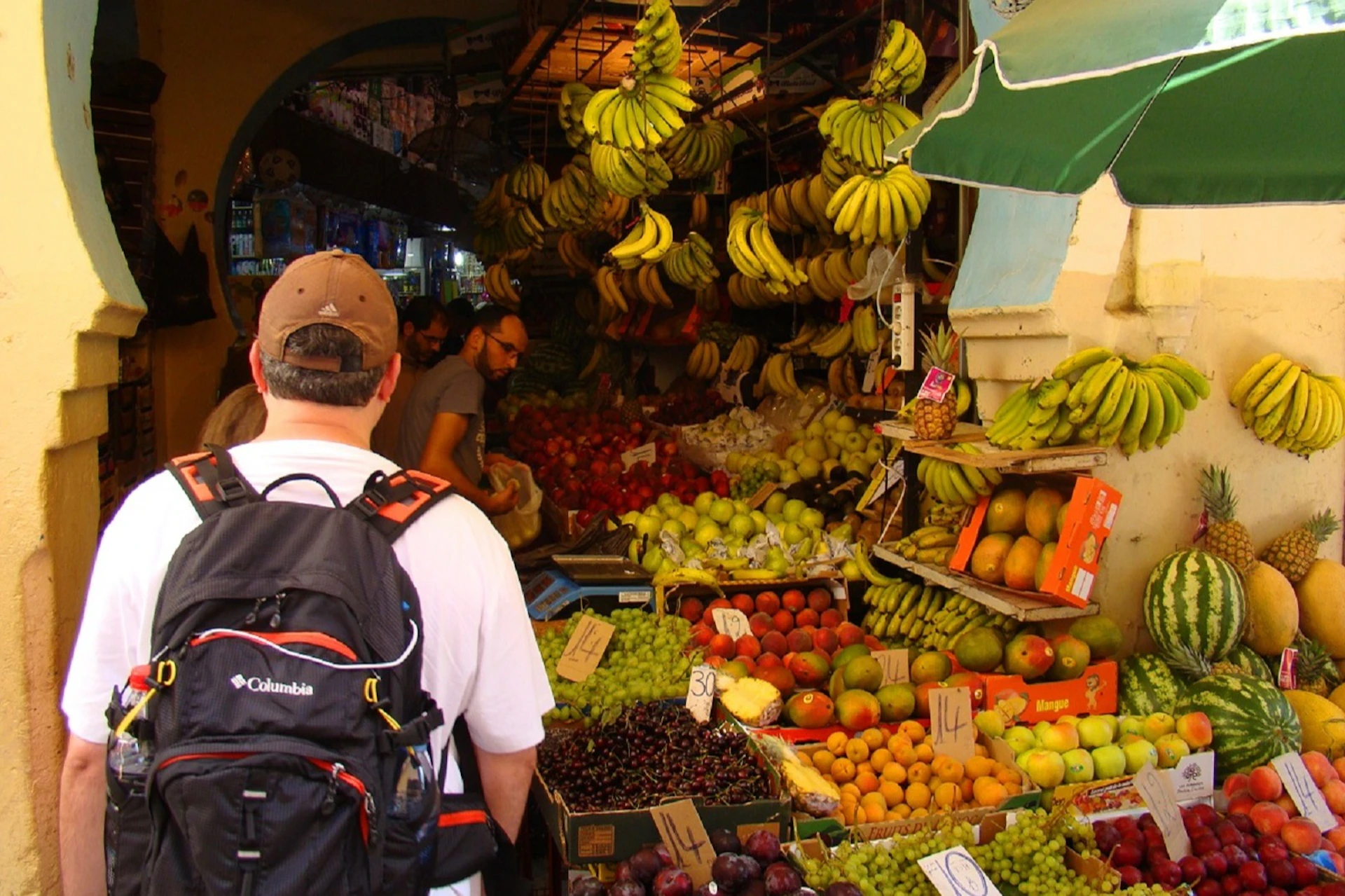 A man with a backpack browses a colorful fruit market, with hanging bananas and various fruits like apples, grapes, and mangoes.