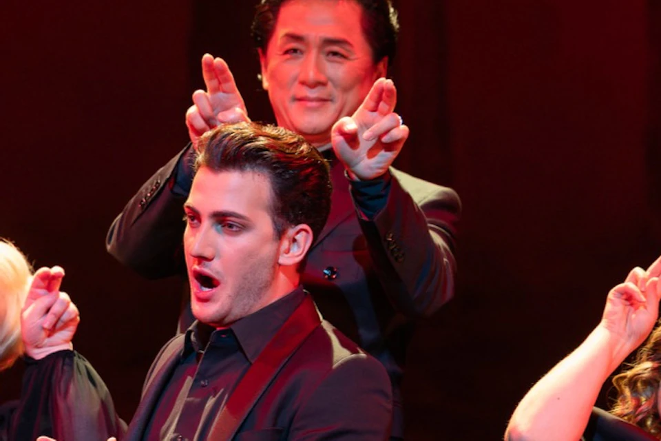 Four performers on stage in black outfits, making playful gestures with their hands, under red stage lighting.