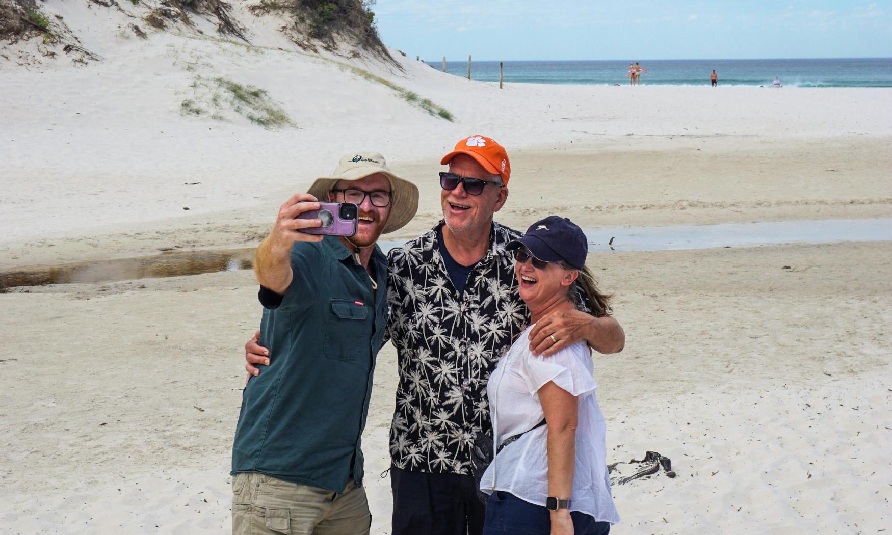 Τρεις άνθρωποι χαμογελούν και βγάζουν μια selfie σε μια αμμώδη παραλία με αμμόλοφους και φόντο τον ωκεανό.