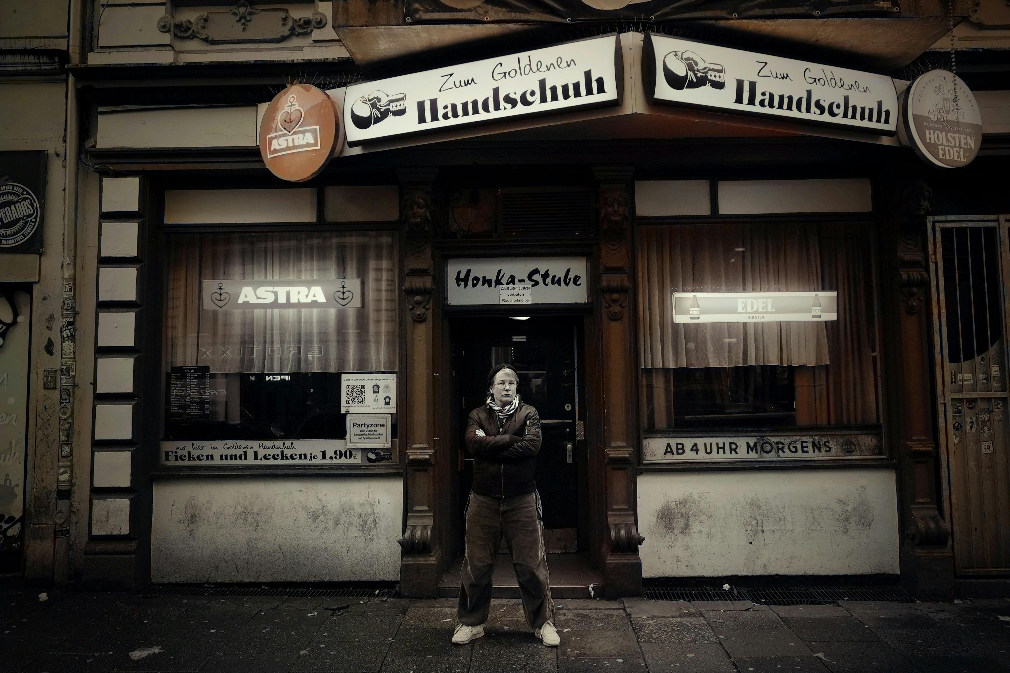 一个人站在名为“Zum Goldenen Handschuh”的昏暗酒吧前，双臂交叉，窗户上挂着标牌和窗帘。