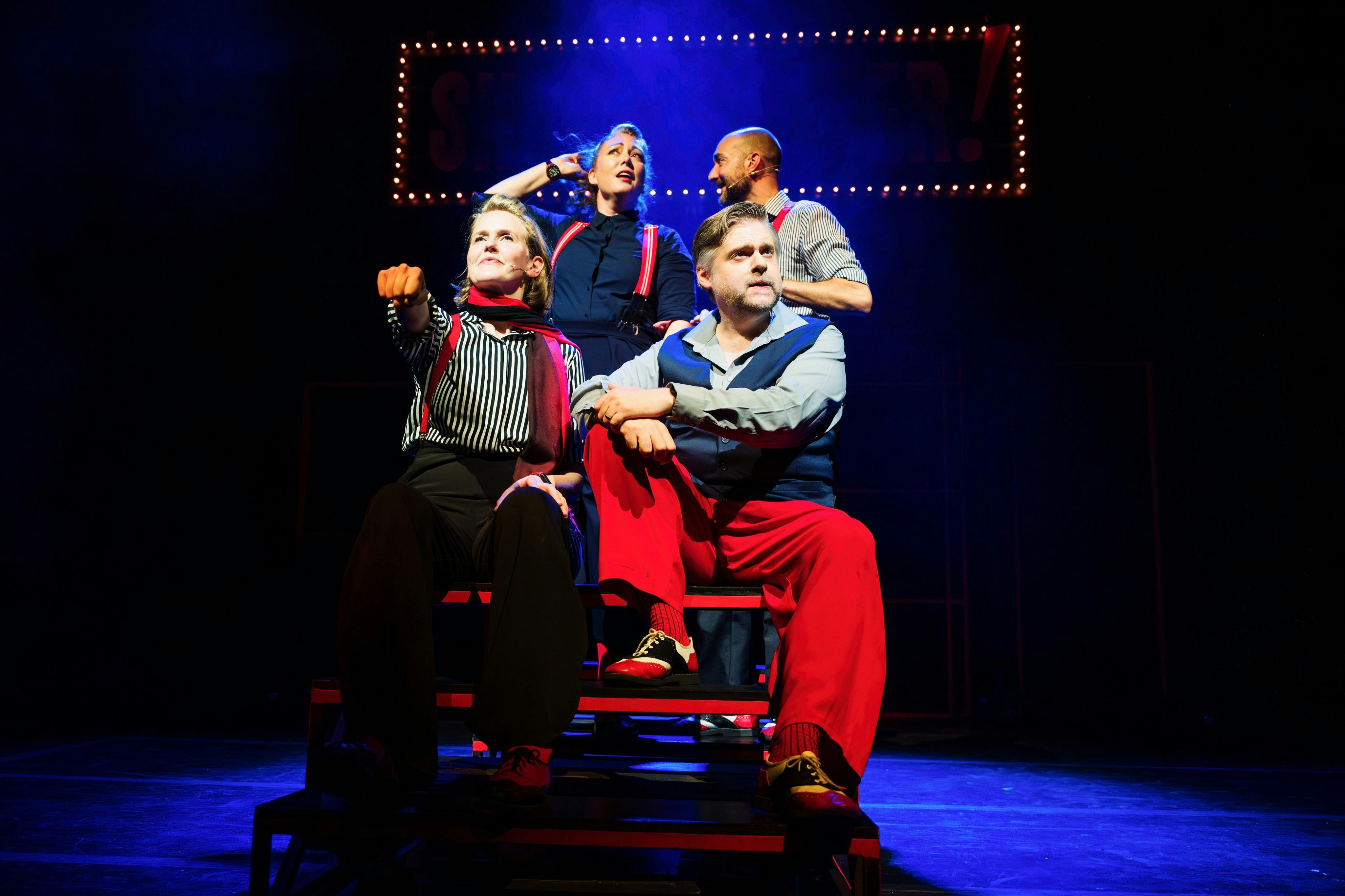 Čtyři lidé v divadelních kostýmech dramaticky pózují na jevišti s osvětleným nápisem v pozadí.