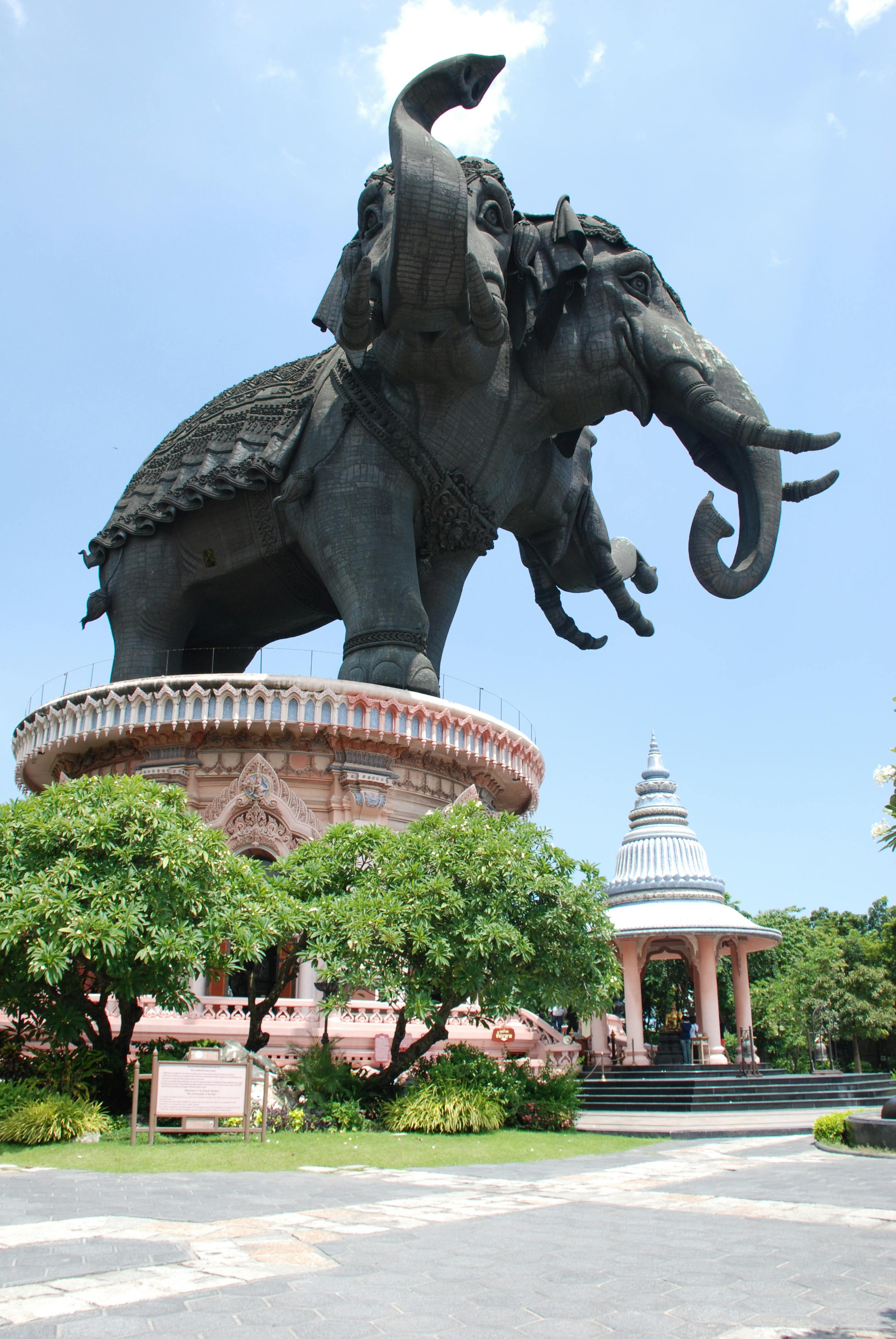 ピンク色の円形のベースにある巨大な3つの頭の象の彫刻。晴れた空の下には、木々と小さな華やかなパビリオンが近くにあります。
